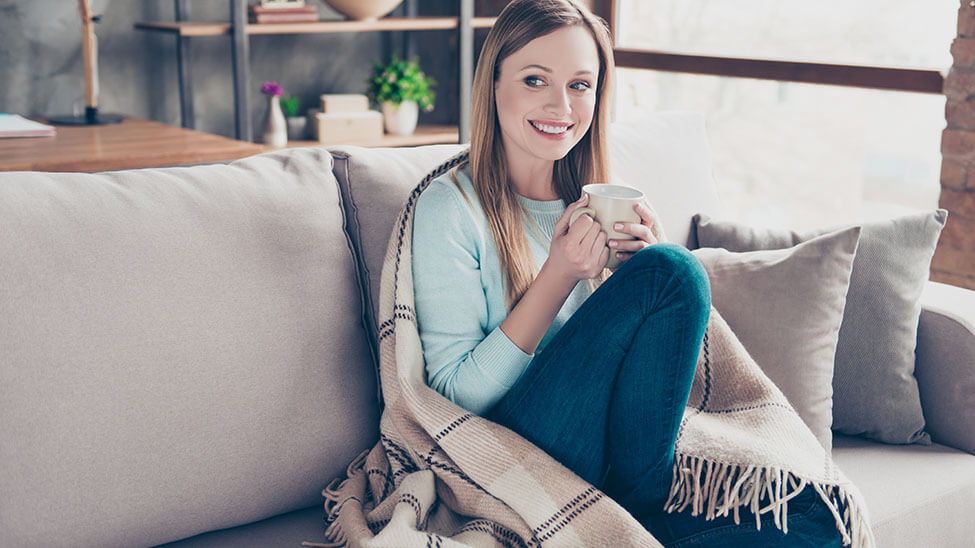 Woman warming herself with blanket and cup of tea at home on sofa