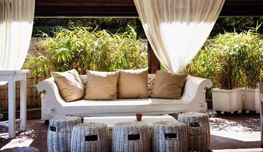 Terrace with big couch, rattan baskets, many bushes in the background and white curtains in front of it