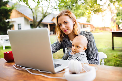Mother sits with baby on lap outside in garden and works on laptop