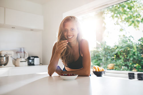 Young woman standing in kitchen smiling and eating fruits