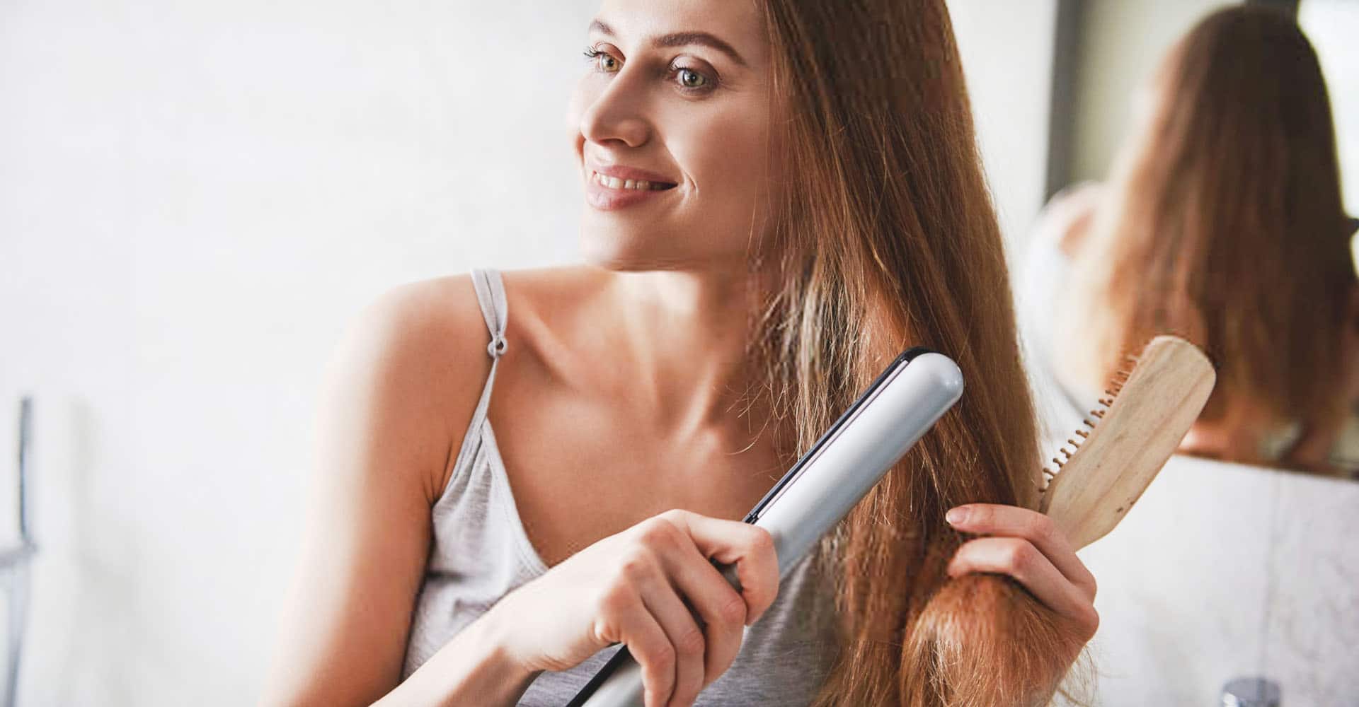 Woman straightening hair in hair straightener in bathroom in front of mirror