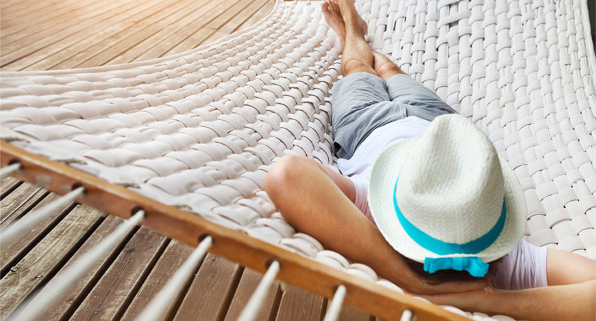 Eine große, weiß geflochtene Hängematte über einem Holzboden, auf der ein Mann mit Hut schläft oder entspannt.