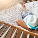 Eine große, weiß geflochtene Hängematte über einem Holzboden, auf der ein Mann mit Hut schläft oder entspannt.