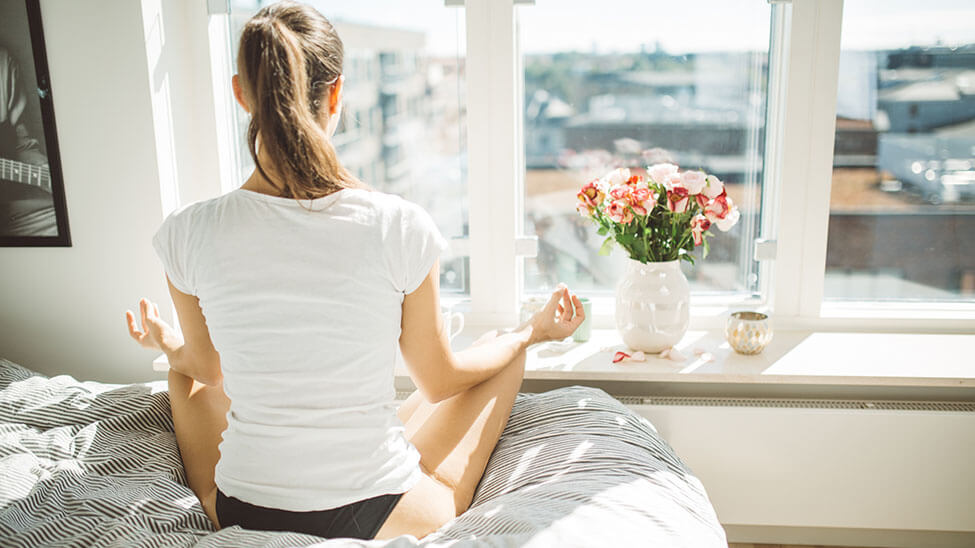 Frau meditiert auf Bett vor Fenster in hellem Zimmer