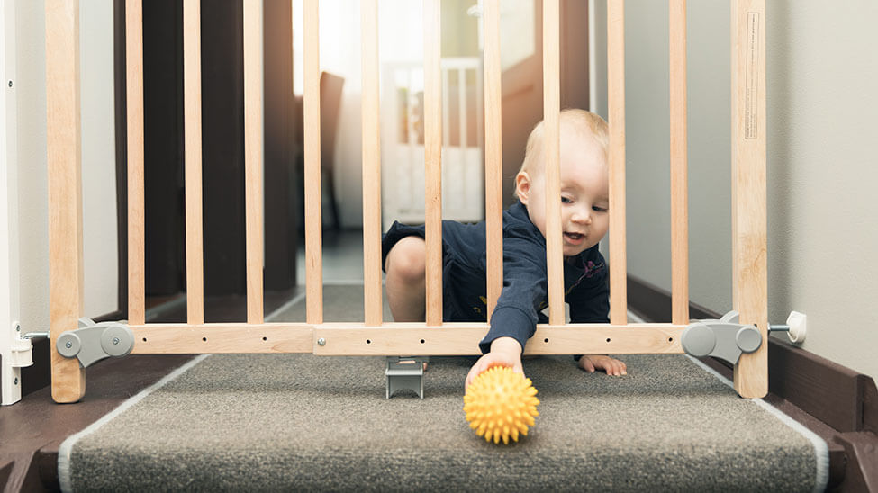 Baby greift durch Schutzgitter nach Ball, der oben auf einer Treppe liegt