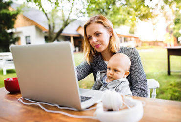 Mutter sitzt mit Baby auf Schoß draußen im Garten und arbeitet am Laptop