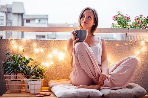 Glückliche Frau auf ihrem Balkon sitzt auf Kissen und trinkt einen Tee