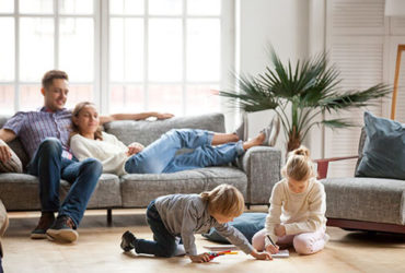 Familie im Wohnzimmer - die Eltern sitzen entspannt auf der Couch, während die Kinder ausgelassen spielen