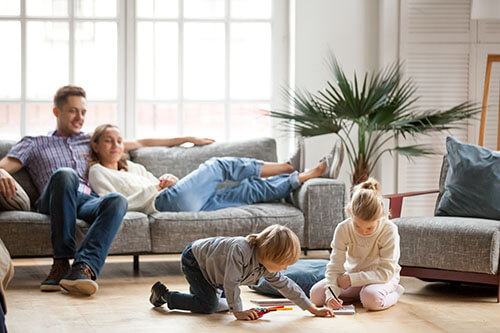 Familie im Wohnzimmer - die Eltern sitzen entspannt auf der Couch, während die Kinder ausgelassen spielen