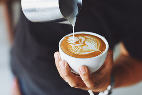 Mann gießt Milch in Kaffee und erzeugt dadurch eine Blume auf dem Kaffee-Schaum