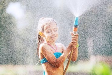 Mädchen kühlt sich im Sommer im Garten mit Wasserschlauch ab
